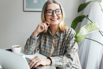 Picture of cheerful female of 60s in casual clothes and glasses enjoying her leisure time using advanced technologies and talking to AI chatbot, discussing funny topics, asking questions