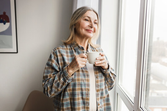 Adorable caucasian smiling senior lady in plaid shirt enjoying her herbal tea standing in cozy room next to big window and admiring first snow and winter coming looking through at city street