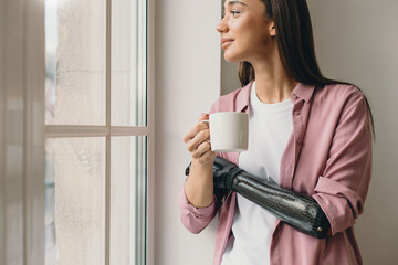 Closeup image of good-looking girl with bionic prosthetic hand holding mug of coffee and looking out the window, craning her neck with curious smiling like she peering at someone outside