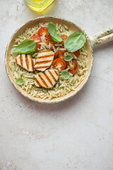 Orzo pasta with grilled halloumi, tomatoes and basil oil in a serving pan, vertical shot on a light-beige stone background with space, above view