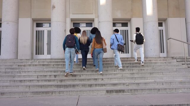 Rear view of a multiracial group of young college friends walking together up the stairs of a university building to attend class