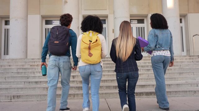 Multiracial group of young friends with backpacks walking up the stairs to the university building. Rear view of college students arriving on campus
