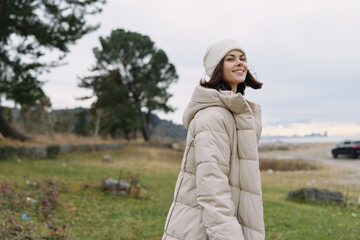 Woman in a light puffer jacket stands in an open field with trees and a distant coast, bright smile and relaxed pose, autumnal nature setting, casual fashion in outdoors