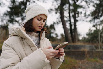 Woman using smartphone outdoors in winter coat and beanie, focused on the device in a park with...