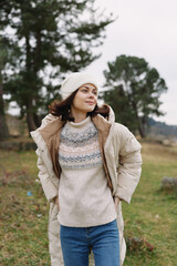 Woman in a cozy winter coat and knit sweater stands outdoors in a grassy field, wearing a hat, with...