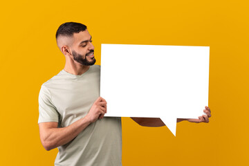 A man stands indoors holding a blank speech bubble sign. He is smiling at the sign, with a yellow background behind him. This setting appears bright and lively.