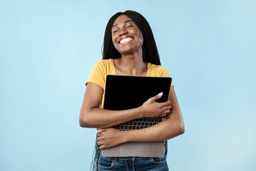 Obraz premium People And Gadgets. Joyful Black Woman Hugging Laptop Standing In Studio Over Blue Background. Lady Posing Holding Computer. Freelance And E-Learning. I Love Internet Technologies Concept