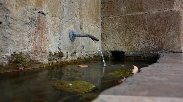 Spain, Navarre, Aranguren, May 24, 2025: An old water fountain embedded in a stone wall. A metal pipe protrudes from the wall and drips a continuous stream of water into a small stone channel or basin