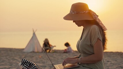 Family enjoying a beach day at sunset with kids playing in the background while one adult works on a laptop