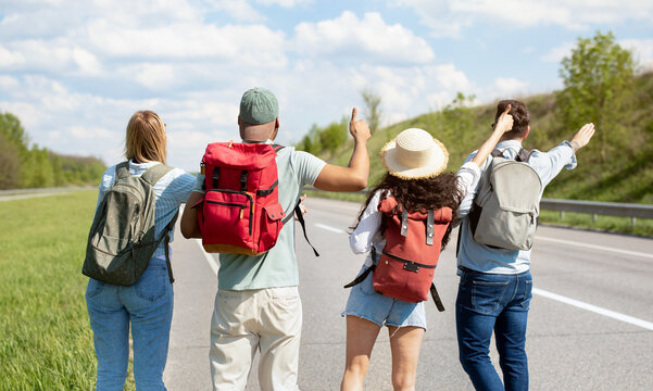 Back view of young multiracial friends standing on highway, showing hitchhiking gesture, waving down car, going on journey together, outdoors. Group of millennial students traveling by autostop