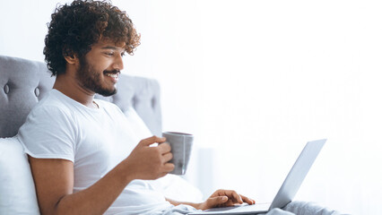 A young man sits on a bed holding a cup of coffee. He is looking at his laptop with a smile in a well-lit room during the day.
