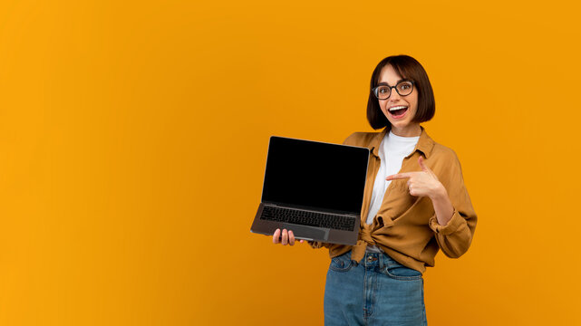 Wow, great website. Excited lady pointing at laptop with black screen, standing over yellow background, panorama with free space. Surprised woman with computer. Mockup image