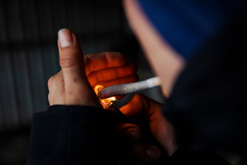 Detailed close-up of hands lighting a hand-rolled marijuana cigarette using a lighter. Flame illuminating fingers in a dark environment. No face clearly visible.