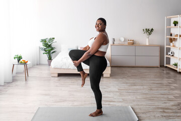 Overweight black lady stretching her leg, making flexibility exercises on yoga mat, leading active...