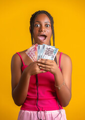 Image of young black African woman with an amazed expression holds a fan of 100 and 200 Ghanaian cedi notes. Set against a yellow backdrop, she wears a pink top and has long braids, capturing a moment
