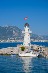 Fototapeta premium Seaside landscape with moored boat and picturesque mountain landscape in background, lighthouse and waving Turkish flag on it.