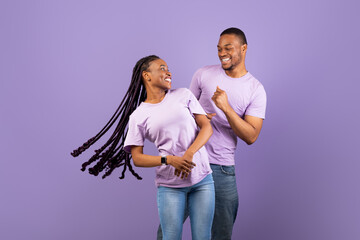 Obraz premium Enjoying Life. Portrait of happy carefree African American partners dancing and having fun, looking at each other, isolated on pastel purple violet studio background, copy space