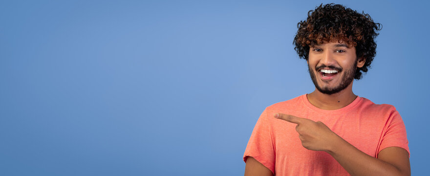 A man stands with a smile on his face and points to the left. He has curly hair and wears a light orange shirt. The background is plain blue.