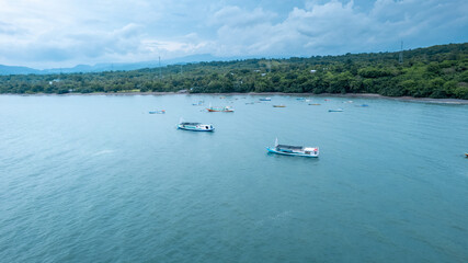 Fototapeta premium High angle landscape of various fishing boats floating on tranquil turquoise sea near a lush green coastline under a cloudy sky in summer morning. 