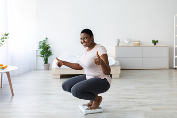 Obraz premium Happy plump African American woman sitting on scales, showing thumb up gesture, reaching her slimming goal, celebrating achievement at home. Successful weigt loss concept