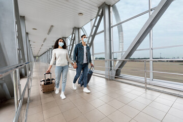 Fototapeta premium Healthcare And Travels. Middle Eastern Couple In Medical Masks Walking With Luggage At Airport Terminal, Young Arab Spouses Holding Hands While Going To Flight, Travelling During Coronavirus Pandemic