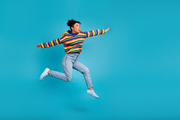 Fototapeta premium Joyful young woman in a colorful striped sweater jumps in mid air against blue background showcasing casual fashion vibrant lifestyle advertising