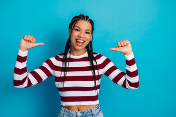 Young woman with braids in striped top smiles confidently and points to herself against a blue...