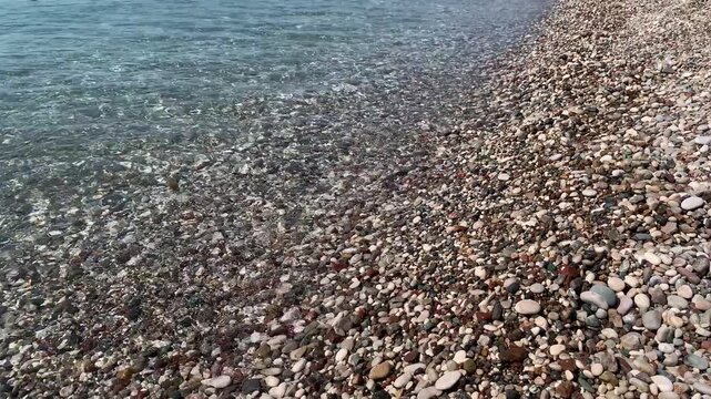 Waves gently wash over colorful pebbles on a sunny day. Enjoy the view of the shoreline and the calm water.