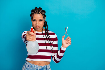 Young woman with striped top holding spray bottle and scissors stands against blue background