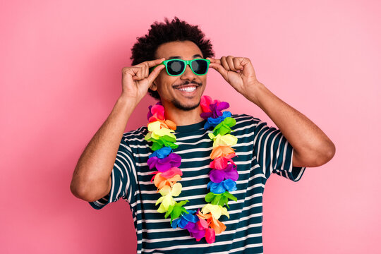 Young mixed race man wearing a rainbow lei and green sunglass smiles against a pink background in a fun casual fashion lifestyle portrait