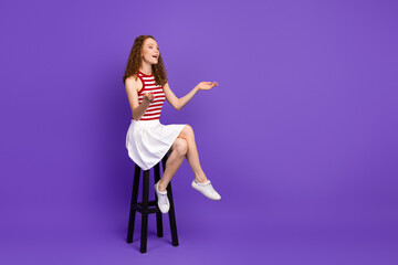 Young woman on purple background smiling and gesturing while sitting on a stool wearing red white...
