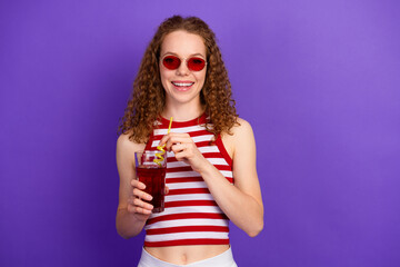 Young woman with striped top and red sunglass smiles while sipping a drink against a purple...
