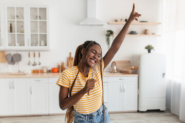Cheerful millennial pretty black female singing at utensil as imaginary microphone, dancing in...