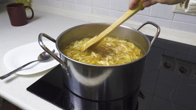 Woman stirs shredded cabbage and diced potatoes in a boiling pot with a wooden spatula. Cooking authentic Ukrainian borscht soup in a home kitchen