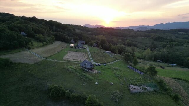 Aerial video of traditional Romanian wooden houses and a greenhouse in Glod village, Alba County, Romania, at golden sunset on August 15, 2025, with rolling hills and mountain horizon.