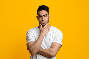 Doubting handsome millennial middle eastern guy in casual outfit touching his chin and looking at...
