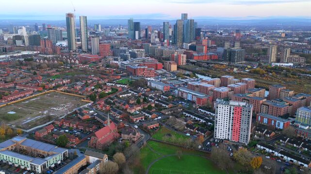 Established aerial video of Ordsall Park in Salford, capturing the local football pitch, the historic red-brick St Clement&rsquo;s Church, and the towering skyscrapers of Manchester city center.