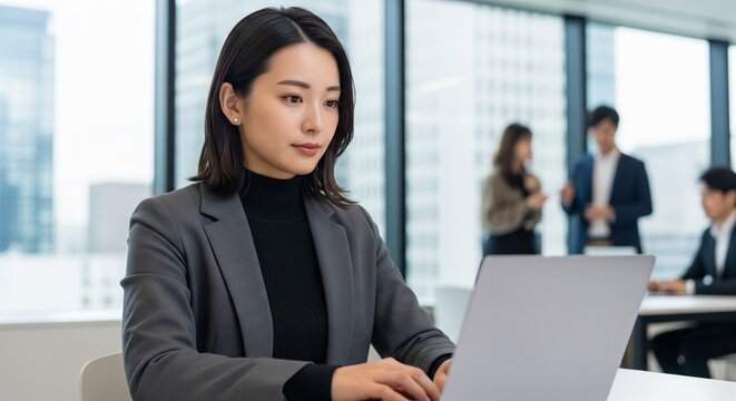 モダンなオフィスでノートパソコンに向かって仕事をするジャケットとタートルネック姿の20代日本人女性社員