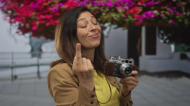 Woman holds vintage camera making middlefinger gesture on street under blooming bougainvillea; defiance rebellion protest resistance.