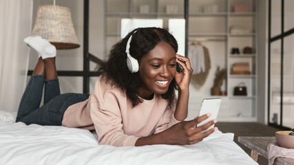 Black Millennial Woman Using Smartphone Making Video Call Wearing Wireless Earphones Lying In Bed In Modern Bedroom At Home. African Lady Listening To Music On Phone Mobile Application. Panorama