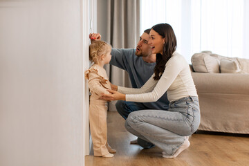 Kids Growth Progress. Loving parents fixing height of sweet little daughter child, setting hand on...