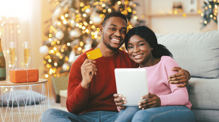 A couple sits on a couch together, smiling while using a tablet for online shopping. The room is decorated for the holidays with lights and gifts around them.