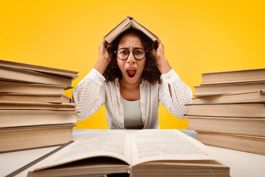A student is sitting at a table surrounded by stacks of books. She holds a book over her head and looks stressed. The background is bright yellow, indicating a study environment.