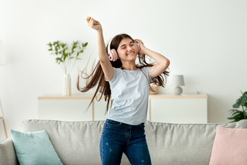 Happy Indian teenage girl with headphones dancing to beautiful music at home. Cheerful adolescent...