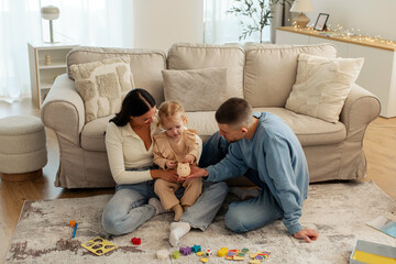 Happy young parents and toddler girl putting cash into piggy bank while playing with child on...