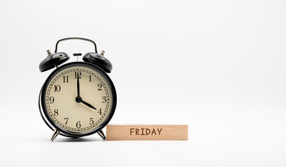 Alarm clock showing 4 o'clock next to wooden block with Friday text on white background. 