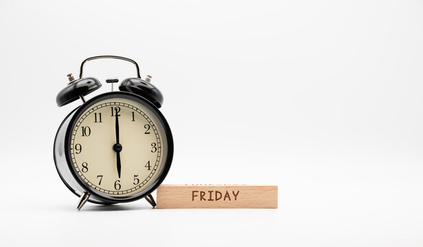 Alarm clock showing 6 o'clock next to wooden block with Friday text on white background. 