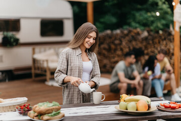 Happy millennial Caucasian woman pouring tea, making breakfast near RV, camping with her diverse...