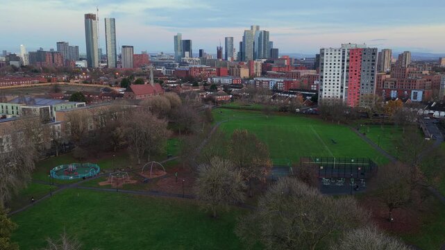 Established aerial video of Ordsall Park in Salford, capturing the local football pitch, the historic red-brick St Clement&rsquo;s Church, and the towering skyscrapers of Manchester city center.