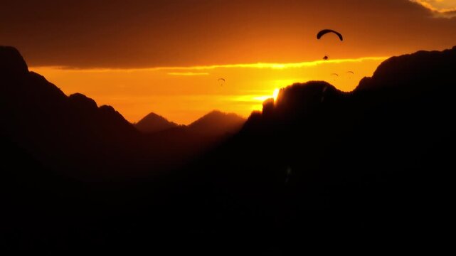 Paraglider flying above dramatic mountain landscape during golden hour at sunset. Scenic aerial view with warm sunlight creating a cinematic and adventurous atmosphere